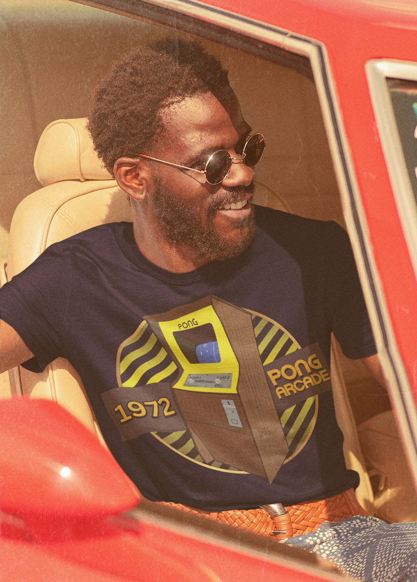 A man sitting in a vintage car, wearing a black T-Shirt with words 1972,pong arcade, image of a pong arcade machine in front of a yellow striped circle