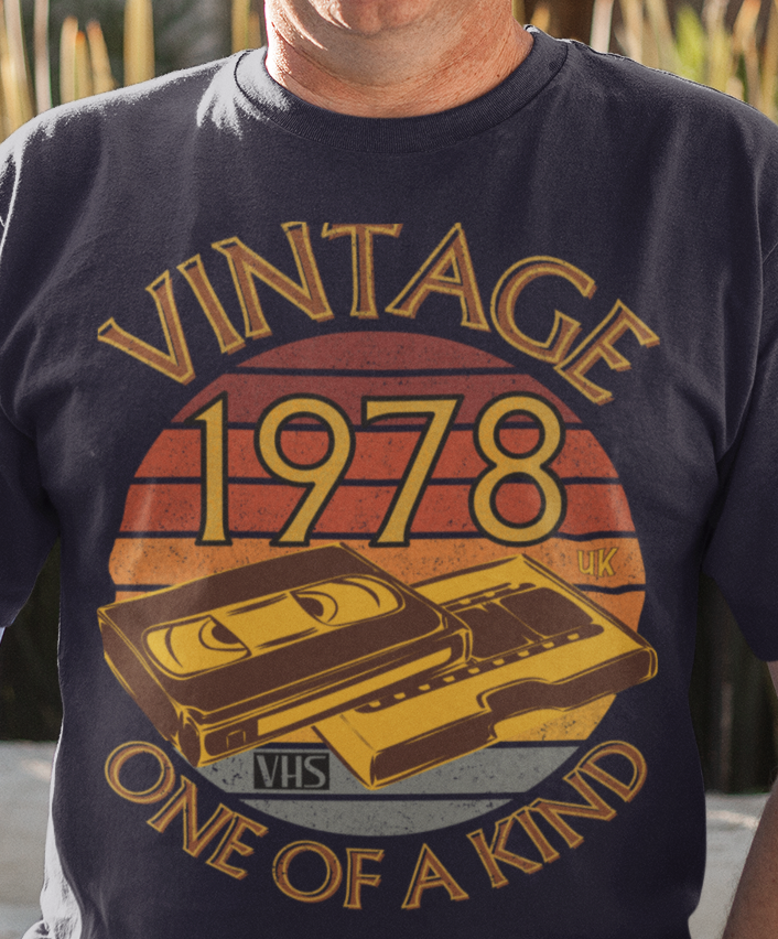 A man wearing a navy T-Shirt, browny sunset circle vintage and one of a kind around it,numbers 1978 UK in  circle & an image of a VHS tape and cover box
