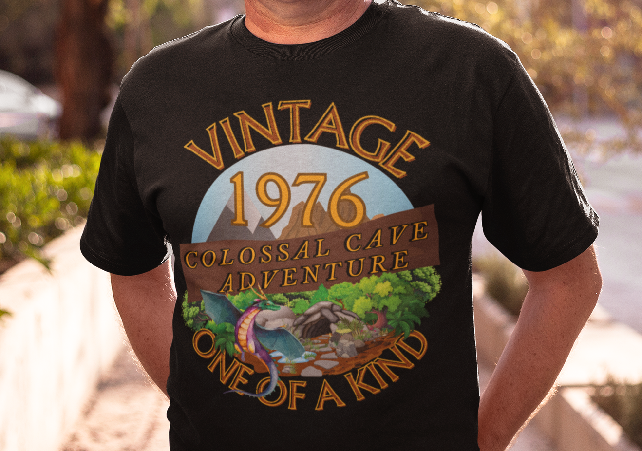 A man wearing a black T-Shirt with words vintage,1976,colossal cave adventure,one of a kind,circular picture of a dragon,cave and woodland,blue skyBlack T-Shirt with words,1976,colossal cave adventure,circular picture of a dragon,cave and woodland,blue sky,daytime